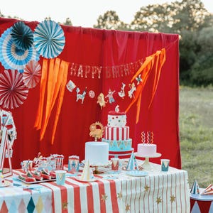 Op de afbeelding: Een feestelijke verjaardagsscene met een rode achtergrond, versierd met papieren waaiers en een "Happy Birthday" banner. Een tafel is gedekt met een tafelkleed met circusthema, taarten en feestartikelen. De sc&egrave;ne speelt zich buiten af, met een grasachtige achtergrond.