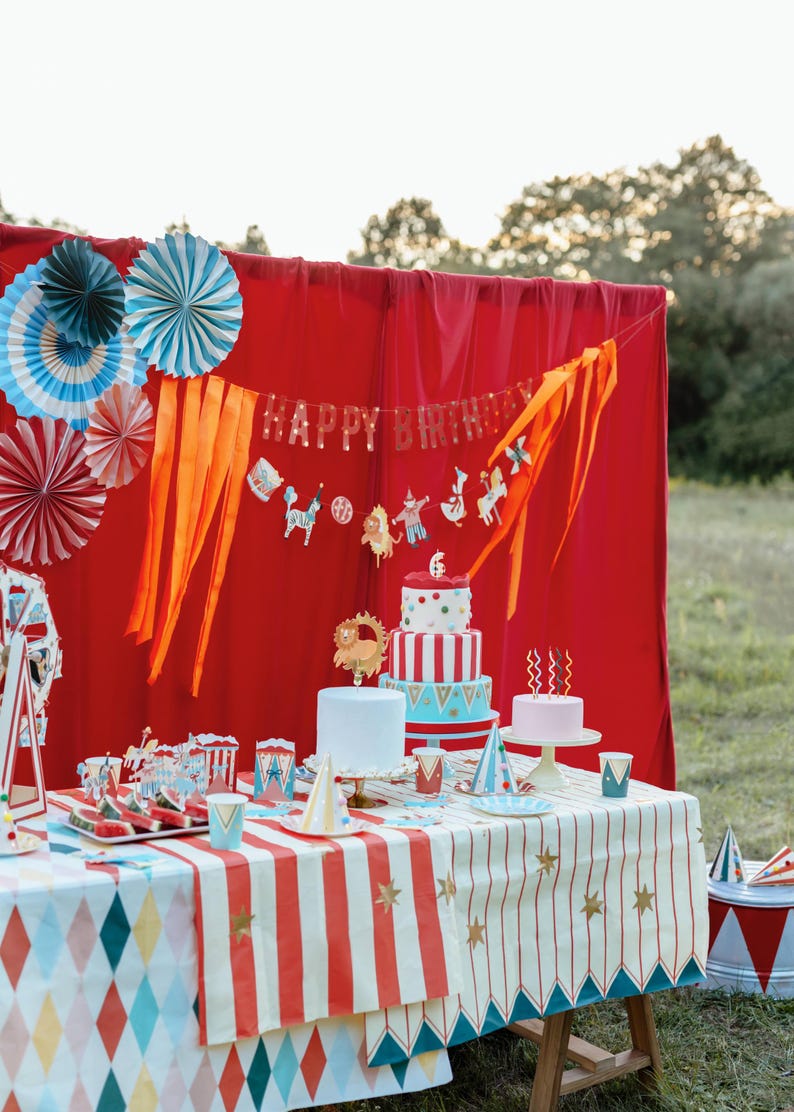 Op de afbeelding: Een feestelijke verjaardagsscene met een rode achtergrond, versierd met papieren waaiers en een "Happy Birthday" banner. Een tafel is gedekt met een tafelkleed met circusthema, taarten en feestartikelen. De sc&egrave;ne speelt zich buiten af, met een grasachtige achtergrond.