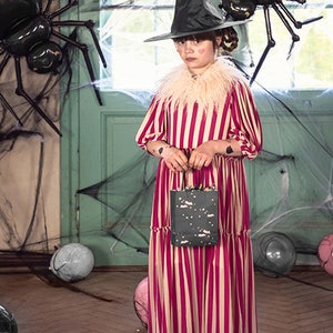 May include: A young person dressed as a witch for Halloween, wearing a black and white striped dress, a black witch hat, and black boots. They are holding a black and white patterned bag. The background features a green door with a spider web and two black spider decorations.