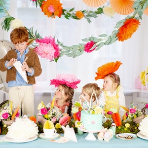 May include: A table setting for an Easter party with a pastel blue tablecloth, white plates, and a white cake decorated with flowers. There are also colorful paper flowers, a chick balloon, and a bunny balloon on the table. Four children are sitting at the table, wearing colorful hats and looking at a white paper cone.