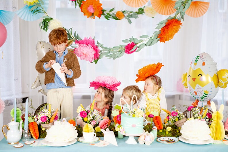 May include: A table setting for an Easter party with a pastel blue tablecloth, white plates, and a white cake decorated with flowers. There are also colorful paper flowers, a chick balloon, and a bunny balloon on the table. Four children are sitting at the table, wearing colorful hats and looking at a white paper cone.