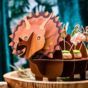May include: A brown cardboard Triceratops dinosaur decoration with a detailed design. The dinosaur has three horns, a frill, and a smiling face. The dinosaur is part of a party decoration set, with other dinosaur-themed food toppers.