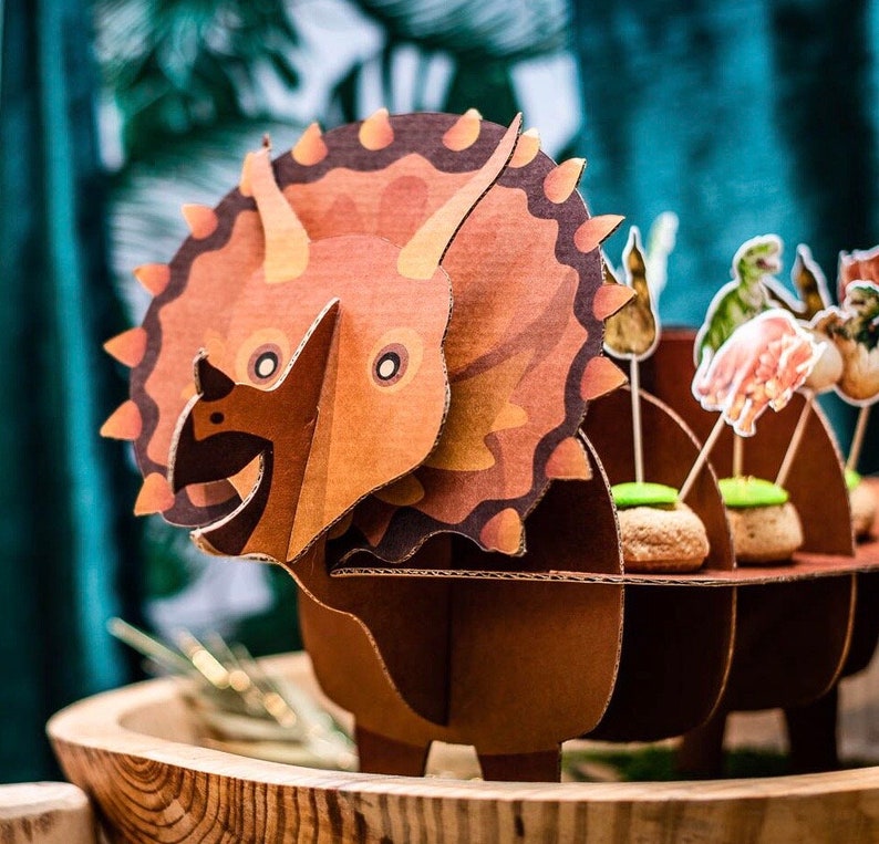 May include: A brown cardboard Triceratops dinosaur decoration with a detailed design. The dinosaur has three horns, a frill, and a smiling face. The dinosaur is part of a party decoration set, with other dinosaur-themed food toppers.