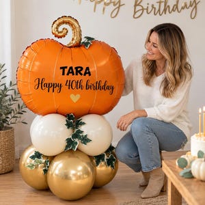 May include: A large orange pumpkin-shaped foil balloon with a gold stem, featuring the black text TARA and Happy 40th birthday with a small gold heart. The balloon sits atop a cluster of white and metallic gold balloons accented with green leaves.