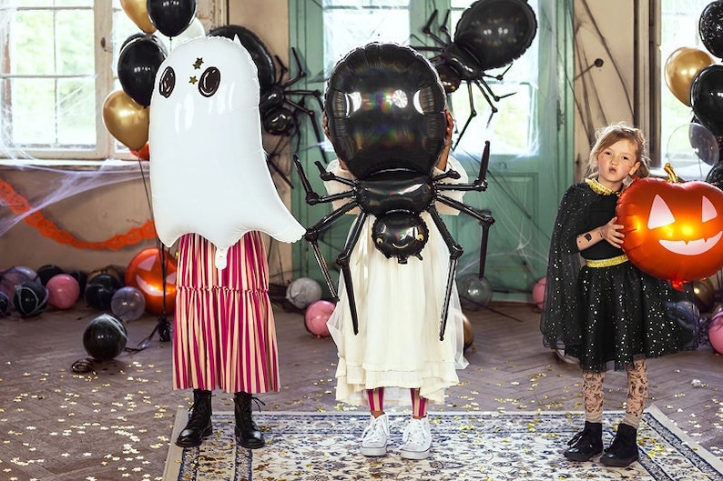 May include: Three children dressed in Halloween costumes holding large inflatable balloons. The child on the left is holding a white ghost balloon. The child in the middle is holding a black spider balloon. The child on the right is holding an orange pumpkin balloon.