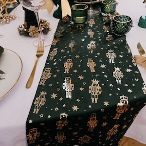 May include: A dark green table runner with a repeating pattern of gold nutcracker figures and stars. The runner is placed on a white tablecloth, with gold cutlery and decorative glassware visible.