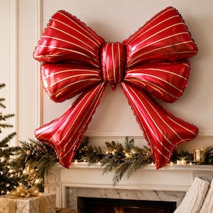 May include: A large, shiny red foil balloon shaped like a decorative bow. The balloon features thin, vertical gold stripes running across its surface, creating a metallic finish. It is designed as a festive wall decoration with two loops and two tails.
