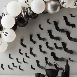 May include: A Halloween-themed display featuring a balloon arch with white, black, and silver balloons, some decorated with ghost and spider designs. Black bat cutouts are affixed to the wall, along with a black cauldron, bottles, and decorative items.