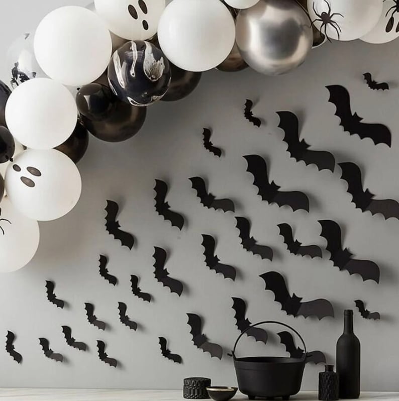 May include: A Halloween-themed display featuring a balloon arch with white, black, and silver balloons, some decorated with ghost and spider designs. Black bat cutouts are affixed to the wall, along with a black cauldron, bottles, and decorative items.