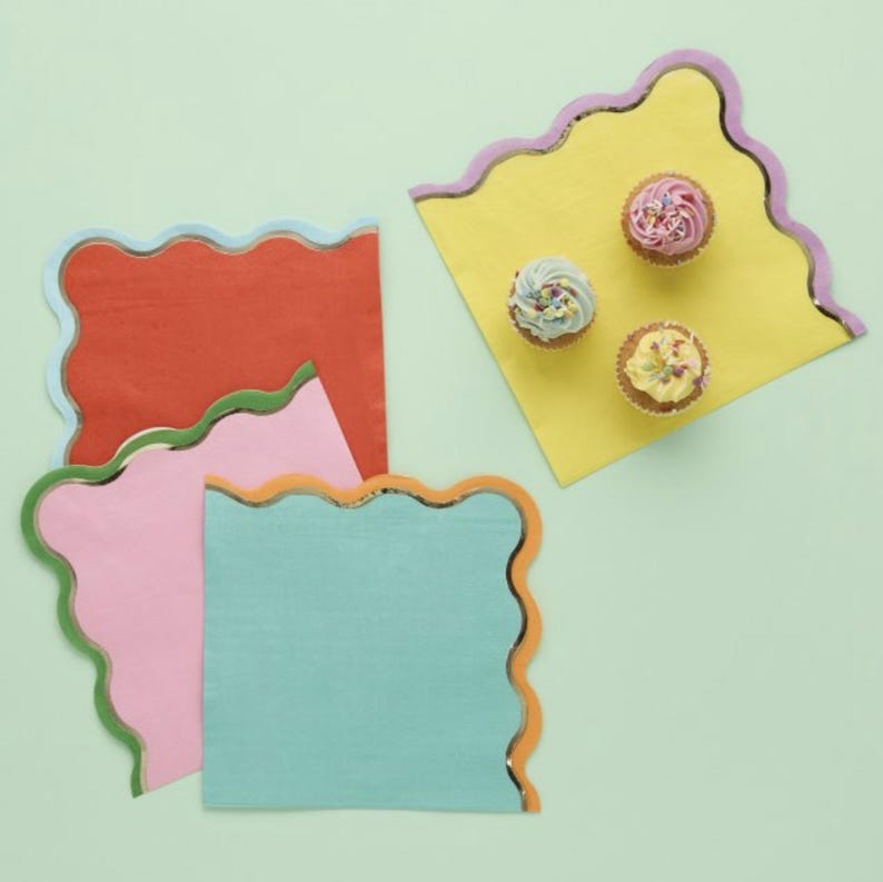 May include: Four colorful paper napkins with scalloped edges and gold foil trim. The napkins are in shades of orange, pink, blue, and yellow. The yellow napkin has three small cupcakes on top.