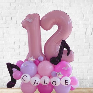May include: A pink balloon arrangement for a birthday celebration. The arrangement includes the number "12" in large pink balloons, smaller pink and purple balloons, and black musical note accents. The name "Chloe" is spelled out in white letter balloons.