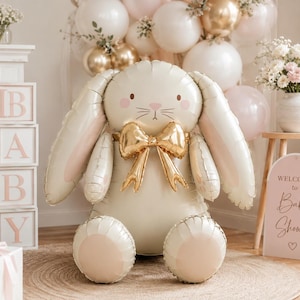Può includere: Un grande palloncino in alluminio color crema a forma di coniglio seduto con orecchie lunghe, un fiocco dorato metallizzato al collo e lineamenti semplici. Il palloncino a forma di coniglio è posizionato al centro, circondato da decorazioni.