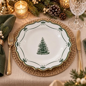 Puede incluir: Un plato de cerámica blanca con borde festoneado, que presenta una ilustración central de un pino verde y un borde decorativo verde con detalles de lazos. El plato se acompaña de un tenedor y un cuchillo de mesa de tono dorado.