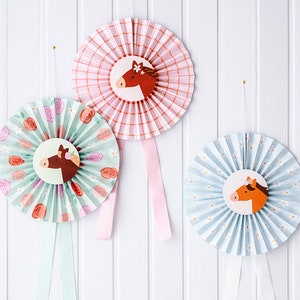 Pode incluir: Três rosetas de papel com desenhos de cabeça de cavalo. As rosetas são xadrez rosa e branco, floral azul e branco e floral verde e branco. Cada roseta tem uma fita rosa, azul ou verde.