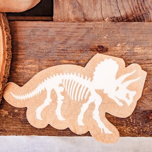 May include: A tan-coloured, die-cut dinosaur skeleton sticker featuring a Triceratops design. The white skeleton graphic is detailed and placed on a light brown background. The sticker is on a wooden surface.