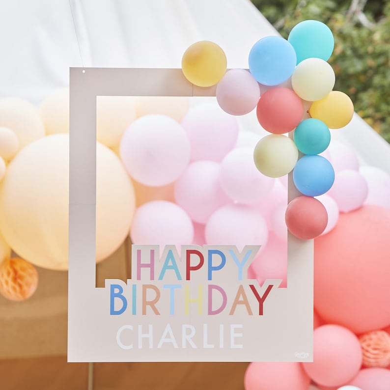 May include: White photo booth frame with the words "Happy Birthday Charlie" in colorful letters. The frame is decorated with a cluster of pastel balloons in pink, yellow, blue, and green.