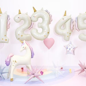 Könnte beinhalten: Dekorationen für eine Party zum Thema Einhörner. Enthält weiße Zahlenballons (1-5), eine Einhornfigur, sternförmige und herzförmige Ballons. Die Ballons haben goldene Akzente und Pastellfarben.