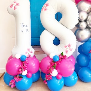 May include: White number 18 balloons with pink floral accents, atop a base of blue, pink, and white balloons. The number 1 has the text "Tara is 18". A festive decoration for an eighteenth birthday celebration.