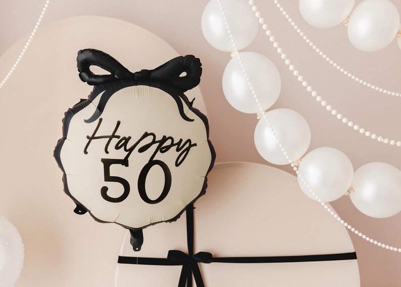 K&ouml;nnte beinhalten: Ein wei&szlig;er Ballon mit einem schwarzen Band und dem Text "Happy 50" in schwarzer Schrift. Der Ballon ist von wei&szlig;en Perlenketten umgeben.