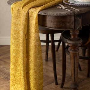 May include: A gold, shimmering table runner draped over a dark wooden table. The table is set with plates, silverware, and a decorative centerpiece. The runner adds a touch of elegance to the dining setting.