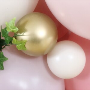 May include: A close-up of a cluster of balloons in pink, white, and gold. The balloons are arranged in a random pattern and are partially obscured by a green leafy vine.
