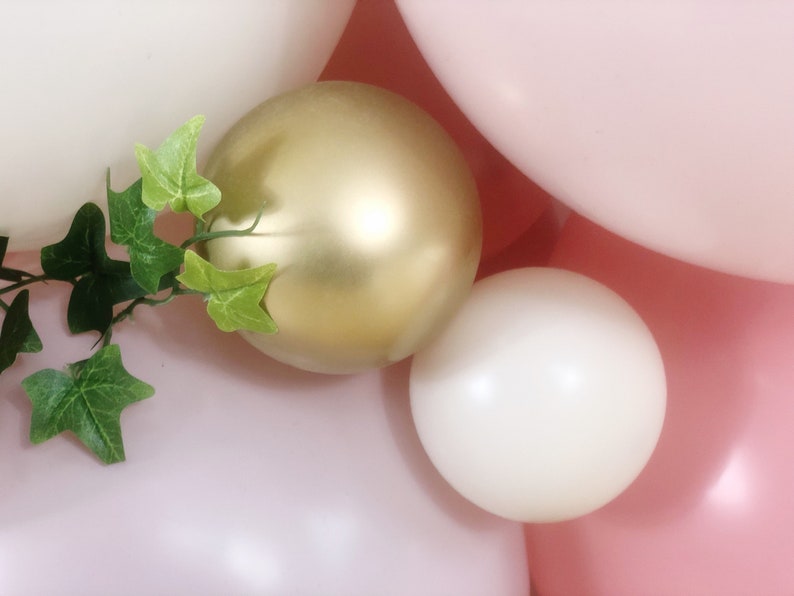 May include: A close-up of a cluster of balloons in pink, white, and gold. The balloons are arranged in a random pattern and are partially obscured by a green leafy vine.