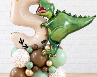 Pode incluir: Um balão grande bege em forma de número cinco com o texto Tara is Five, acompanhado por um balão de dinossauro verde na lateral. A base consiste num conjunto de balões redondos em tons de castanho, verde sálvia, dourado e branco com padrão leopardo.