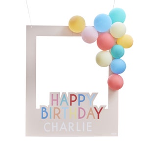 May include: A beige photo booth frame with the words "Happy Birthday Charlie" in colorful letters. The frame is decorated with a cluster of pastel balloons in various colors, including blue, yellow, pink, and peach.