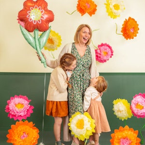 May include: A woman and two children stand in front of a wall decorated with paper flowers. The woman is wearing a floral dress and smiling. The children are wearing casual clothing and looking at the camera. The flowers are in various colors, including pink, yellow, and orange.