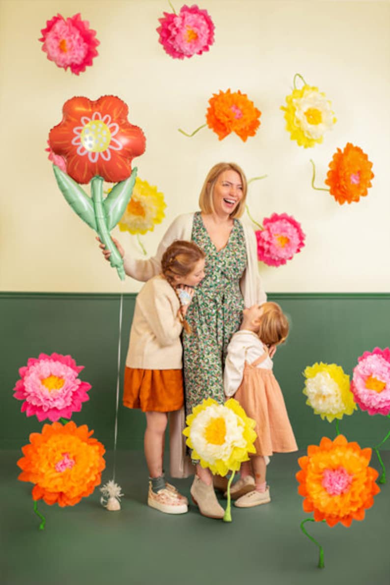May include: A woman and two children stand in front of a wall decorated with paper flowers. The woman is wearing a floral dress and smiling. The children are wearing casual clothing and looking at the camera. The flowers are in various colors, including pink, yellow, and orange.