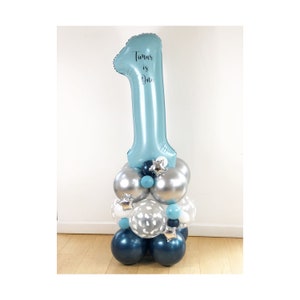 May include: A blue number one balloon with the text "Timur is One" on it. The balloon is on top of a balloon cluster with silver, blue, and white balloons.