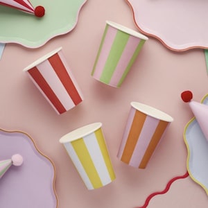 May include: Four striped paper cups in different colors: red and white, green and white, orange and white, and yellow and white. The cups are on a pink background.