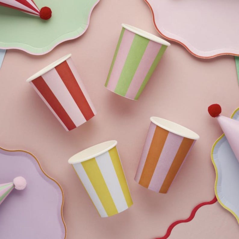 May include: Four striped paper cups in different colors: red and white, green and white, orange and white, and yellow and white. The cups are on a pink background.