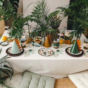 May include: A table setting for a dinosaur-themed party with green and brown paper plates, cups, and napkins. The table is decorated with green plants and a pineapple. There are also green and brown party hats on the table.
