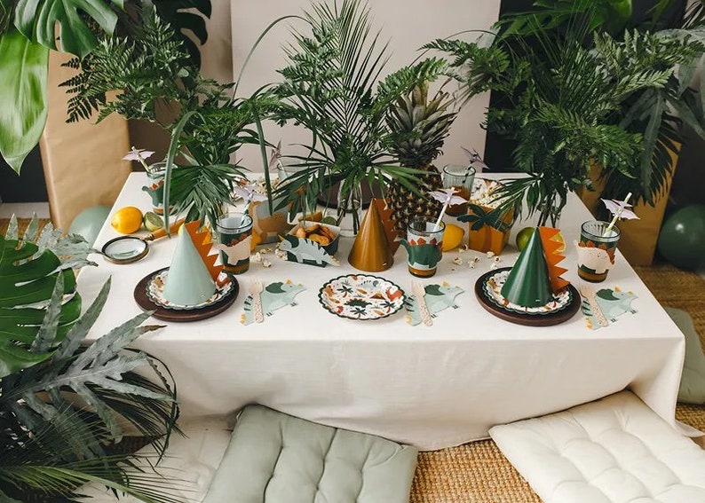 May include: A table setting for a dinosaur-themed party with green and brown paper plates, cups, and napkins. The table is decorated with green plants and a pineapple. There are also green and brown party hats on the table.