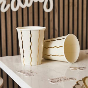Peut inclure: Deux gobelets en papier blanc avec des rayures ondulées dorées, un debout et un sur le côté. Les gobelets sont posés sur une surface blanche avec des découpes décoratives qui disent "joyeux anniversaire". L'arrière-plan est un mur en bois.