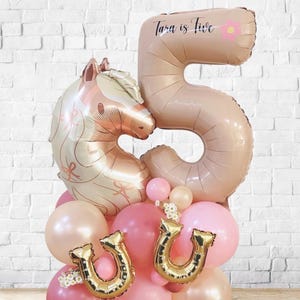 May include: A festive balloon arrangement featuring a large number five balloon, a horse-shaped balloon, and several smaller balloons in shades of pink and gold. The number five balloon has the text "Tara is five". Gold horseshoe balloons add a celebratory touch.