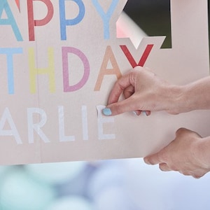 May include: A pink and white cardboard sign with the words "Happy Birthday Arlie" in colorful letters. A person is shown attaching the letters to the sign.