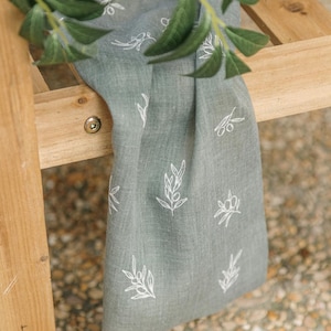 May include: A sage green linen table runner with a white olive branch pattern. The runner is draped over a wooden surface, with fresh green olive branches in the foreground. The fabric has a textured appearance.