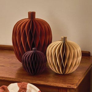 May include: Three pumpkin-shaped decorations made of folded paper with a honeycomb structure. The largest is rust-coloured, the medium is light tan, and the smallest is a deep plum purple. Each features a small, vertical, pleated paper stem.