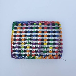 May include: A rainbow-colored woven wallet made from recycled aluminum pull tabs. The wallet is woven with black string and has a black fabric backing.