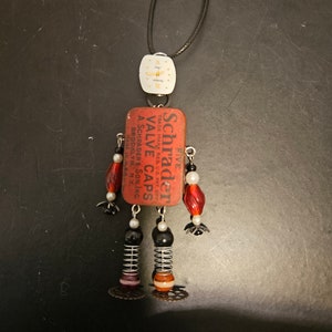 May include: A red and black robot-shaped necklace with a vintage Schrader Valve Caps tin, a watch face, and black beads.