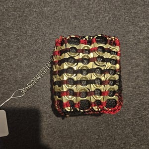 May include: A handmade wallet crafted from repurposed gold-colored can tabs, black material, and red and black thread. The wallet has a silver chain and a white tag attached.