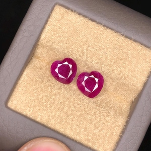 May include: Two heart-shaped, faceted, deep red gemstones. The gems are displayed on a beige, textured surface, set within a gray case. The gems have a bright, reflective quality, showcasing their cut.