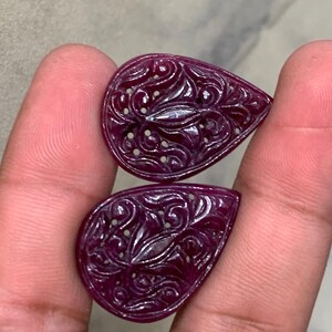 Puede incluir: Dos piedras preciosas en forma de lágrima de color magenta intenso con intrincados patrones florales tallados en sus superficies. Cada piedra muestra un acabado pulido con recortes visibles dentro de los detallados relieves.