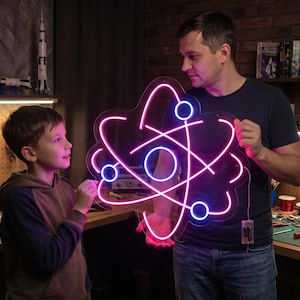 May include: A neon atom sign with pink and blue lights is held by a man and a child. The sign is the focal point, with a workshop setting in the background, including tools and a model rocket.