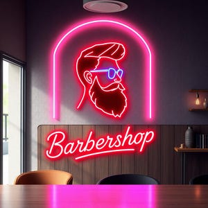 Puede incluir: Un letrero de neón rosa con un contorno rojo de la cara de un hombre que lleva gafas azules y barba. El letrero dice "Barbershop" en letras de neón rojas.