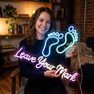 May include: A custom neon sign featuring the text "Leave Your Mark" in a cursive font with two glowing footprints above the words. The sign emits a bright light with blue footprints and purple lettering, connected by a thin white power cord.