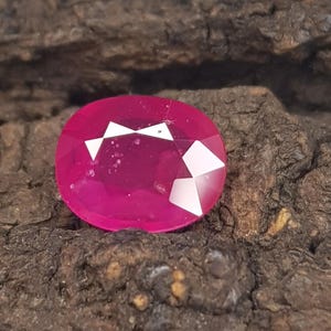 Può includere: Una gemma dal taglio ovale vibrante con una tonalità rosa magenta intenso. La pietra presenta molteplici sfaccettature triangolari e trapezoidali che riflettono la luce, creando luminosi riflessi bianchi sulla sua superficie lucida e traslucida.