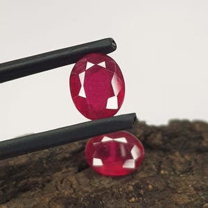 Op de afbeelding: Twee gefacetteerde, ovaalvormige robijnen. Eén robijn wordt vastgehouden door een zwarte pincet, de andere rust eronder op een houten oppervlak. De edelstenen zijn dieprood met witte facetten.